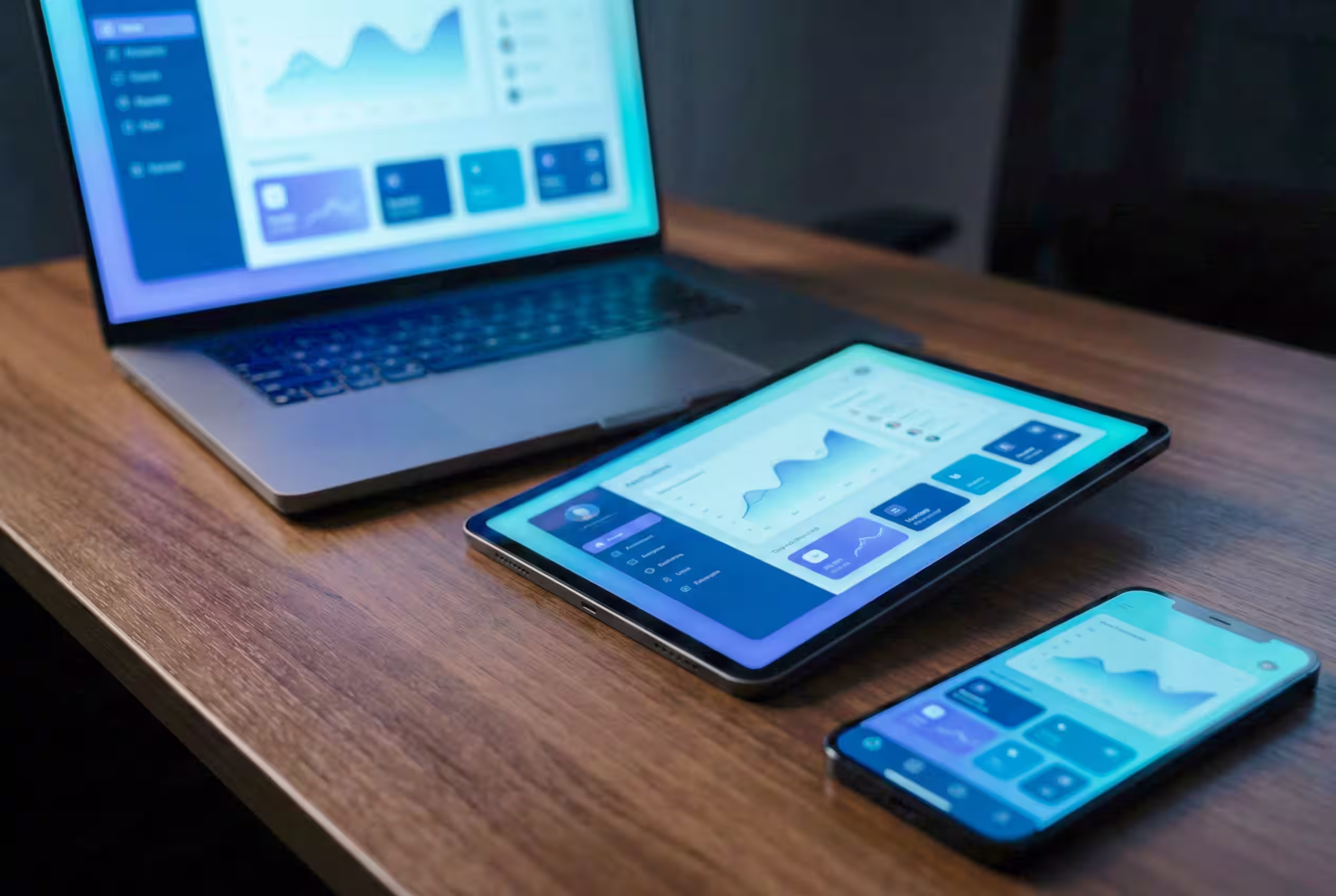 Laptop, tablet, and smartphone on a wooden desk displaying no-code application data charts on their screens built with no-code solutions like Webflow, Airtable, Outseta and Make.